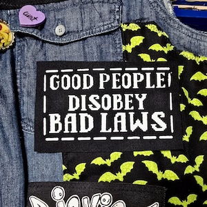 Może przedstawiać: Czarna łatka z białym tekstem, który brzmi "GOOD PEOPLE DISOBEY BAD LAWS". Łatka jest przyszyta do dżinsowej kurtki z zielono-czarną tkaniną w wzór nietoperza.