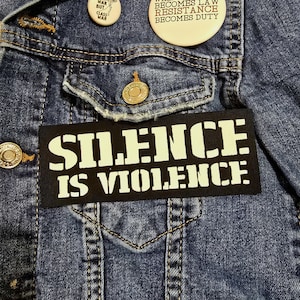 Peut inclure: Gros plan sur une veste en jean bleue avec un patch noir sur lequel on peut lire "SILENCE IS VIOLENCE" en lettres blanches. Deux épingles rondes sont visibles, l'une avec le texte "WAR BUT CLASS WAR" et l'autre avec le texte "WHEN LAW BECOMES RESISTANCE BECOMES DUTY".