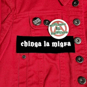 May include: A close-up of a red denim jacket with black buttons. The jacket has three pins: one with the text "Friendly Neighborhood Anarchist", another with the text "Existence Is Resistance", and a black patch with the text "chinga la migra".