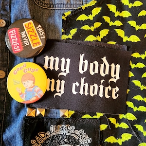 Może przedstawiać: Kurtka dżinsowa z naszywkami. Jedna naszywka jest czarna z białym tekstem, który brzmi "my body my choice". Inna naszywka jest żółta z czarnymi nietoperzami. Trzecia naszywka jest czarna z białym tekstem, który brzmi "SAD QUEERS CLUB".