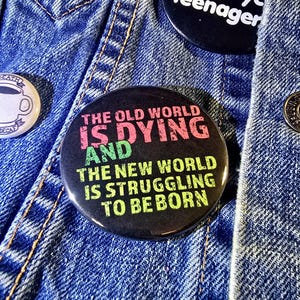 May include: A black button with the text "The old world is dying and the new world is struggling to be born" in neon pink, green, and blue lettering.