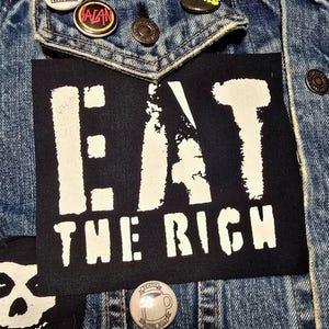 May include: Close-up of a denim jacket with patches and pins. A black patch with the words "EAT THE RICH" in white block letters is centered. Other pins include a round "ALAN" pin and a Misfits skull patch.