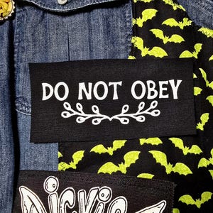 May include: A black patch with white text that reads "DO NOT OBEY" with a white floral design underneath. The patch is sewn onto a denim jacket with a black and green fabric with a bat pattern underneath.