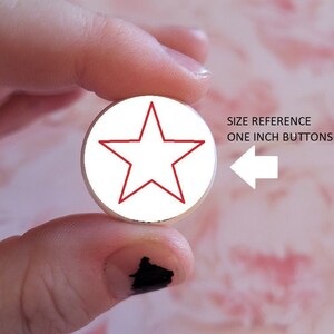 K&ouml;nnte beinhalten: Ein wei&szlig;er Knopf mit einem roten Stern in der Mitte. Der Knopf wird von einem Finger und Daumen gehalten. Der Text "SIZE REFERENCE ONE INCH BUTTONS" ist im Hintergrund sichtbar.