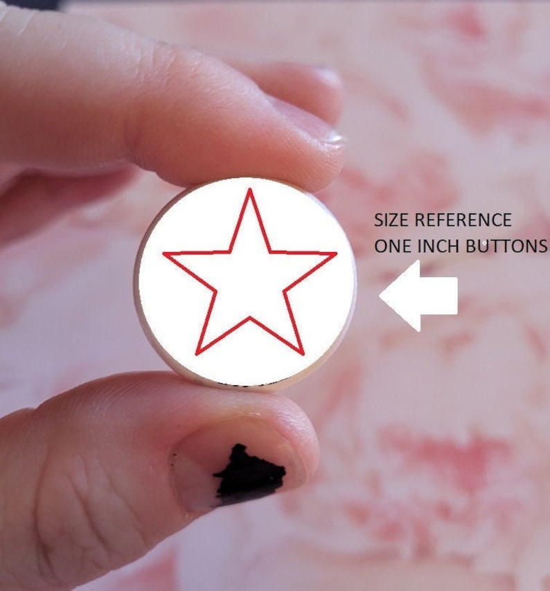K&ouml;nnte beinhalten: Ein wei&szlig;er Knopf mit einem roten Stern in der Mitte. Der Knopf wird von einem Finger und Daumen gehalten. Der Text "SIZE REFERENCE ONE INCH BUTTONS" ist im Hintergrund sichtbar.