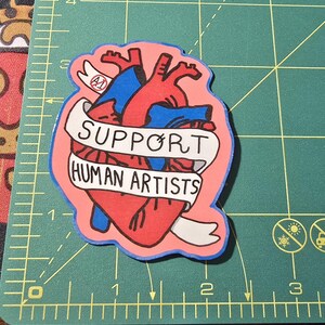 Op de afbeelding: Een sticker met een gestileerd anatomisch hart in rood en blauw, met een banner met de tekst "SUPPORT HUMAN ARTISTS" in zwarte letters. De sticker heeft een roze achtergrond en een blauwe omtrek.