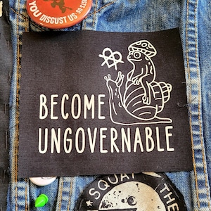 May include: Black patch with white text that reads "Become Ungovernable". A white line drawing of a snail with a mushroom on its back and a creature sitting on top of the mushroom.