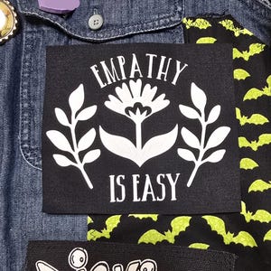 May include: Black fabric patch with white text and a white floral design. The text reads "Empathy is Easy".