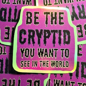 Puede incluir: Una pegatina holográfica con fondo rosa y borde verde. La pegatina presenta texto negro que gotea y dice "Sé el críptido que quieres ver en el mundo."