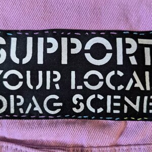 May include: A black patch with white text that reads "SUPPORT YOUR LOCAL DRAG SCENE" sewn onto a light purple fabric background. The patch is outlined with a multi-colored thread.
