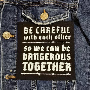Op de afbeelding: Close-up van een blauw spijkerjack met een zwart patch. De patch heeft witte tekst: "BE CAREFUL with each other SO WE CAN BE DANGEROUS TOGETHER". De jas heeft goudkleurige knopen.