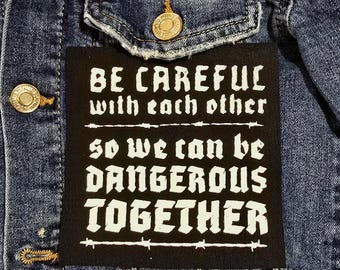 May include: Close-up of a blue denim jacket with a black patch. The patch features white text that reads "BE CAREFUL with each other SO WE CAN BE DANGEROUS TOGETHER". The jacket has gold-colored buttons.
