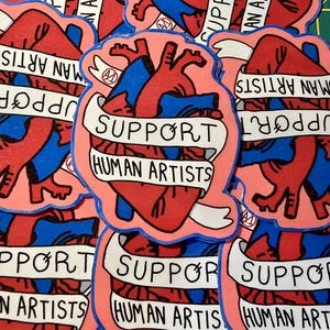 Op de afbeelding: Een verzameling stickers met een gestileerd anatomisch hartontwerp. Het hart is rood en blauw, omlijnd in zwart, met een banner met de tekst "SUPPORT HUMAN ARTISTS". De stickers hebben een roze achtergrond.
