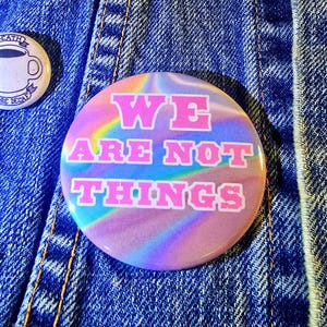 以下が含まれることがあります： ピンクと虹色のボタンに、白い文字で「WE ARE NOT THINGS」と書いてあります。文字の周りはピンクで縁取られています。