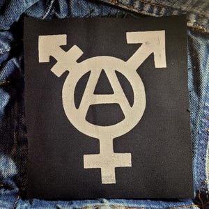 May include: A black fabric patch featuring a white anarchist symbol combined with a transgender symbol. The design includes an encircled letter "A" and gender symbols with arrows. The patch is set against a denim background.