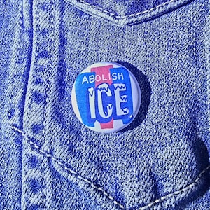 May include: A round button with the words "ABOLISH ICE" in blue and white, with a pink stripe, against a blue denim background. The button is pinned to a denim jacket pocket.