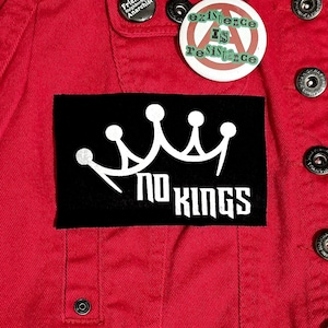 May include: A red denim jacket with several buttons and patches. Patches include a black rectangle with a white crown and the words "NO KINGS", and two round pins with text. The jacket has a classic design.