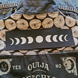 May include: A black denim jacket with a black patch featuring a white crescent moon design. The jacket has a black fabric lining with a repeating pattern of eyes. The lining also has a black patch with the text "OUIJA MYSTIFYING ORACLE" and the words "YES" and "NO".