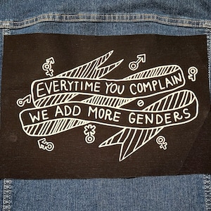 May include: Black fabric patch with white text and illustrations. The text reads "EVERYTIME YOU COMPLAIN WE ADD MORE GENDERS".