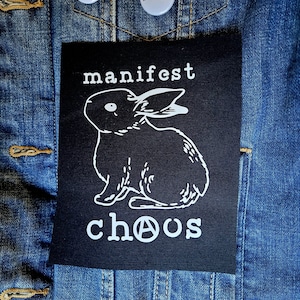 May include: Black and white iron-on patch with a white rabbit and the text "manifest chaos" with an anarchy symbol in place of the "o".