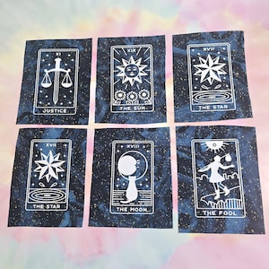 Może przedstawiać: Sześć kart tarota z niebieskim tłem i białymi ilustracjami. Karty są oznaczone cyframi rzymskimi i nazwami kart: Sprawiedliwość, Słońce, Gwiazda, Gwiazda, Księżyc i Głupiec.