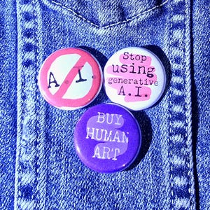 Peut inclure: Trois badges ronds sur un fond en jean. L'un affiche un symbole d'interdiction rouge sur "A.I.". Un autre dit "Stop using generative A.I." en rose. Le troisième badge dit "Buy Human Art" en blanc sur fond violet.
