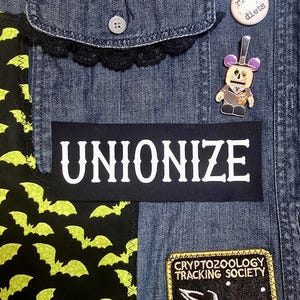 May include: A black denim jacket with a variety of patches, including a black patch with white text that reads "UNIONIZE", a yellow patch with black text that reads "CRYPTOZOOLOGY TRACKING SOCIETY", and a green and black fabric with a pattern of bats.