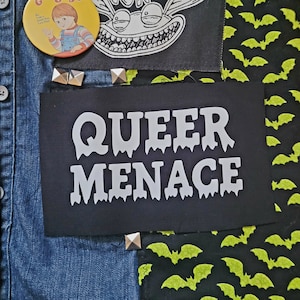 Può includere: Una giacca di jeans nera con varie toppe, tra cui una toppa con il testo "QUEER MENACE" in lettere bianche che gocciolano.
