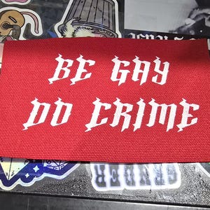 Puede incluir: Un parche rectangular rojo con las palabras "BE GAY DO CRIME" en letras góticas blancas. El parche está sobre una superficie cubierta de pegatinas.