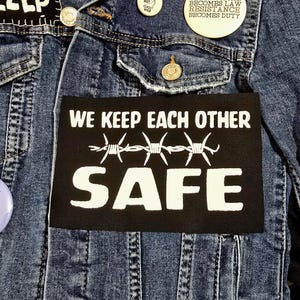 May include: Close-up of a denim jacket with patches and pins. A black rectangular patch with white text reads "WE KEEP EACH OTHER SAFE" above a barbed wire design. Additional pins include text like "WHEN INJUSTICE BECOMES LAW RESISTANCE BECOMES DUTY".
