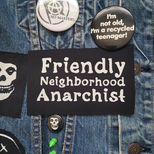 K&ouml;nnte beinhalten: Eine Jeansjacke mit verschiedenen Patches und Pins. Die Patches beinhalten einen schwarz-wei&szlig;en Totenkopf, einen schwarzen Patch mit wei&szlig;em Text, der "Friendly Neighborhood Anarchist" lautet, und einen schwarz-wei&szlig;en Pin mit einem Anarchiesymbol und dem Text "No Gods No Masters". Es gibt auch einen schwarzen Pin mit wei&szlig;em Text, der "Ich bin nicht alt, ich bin ein recycelter Teenager!" lautet.