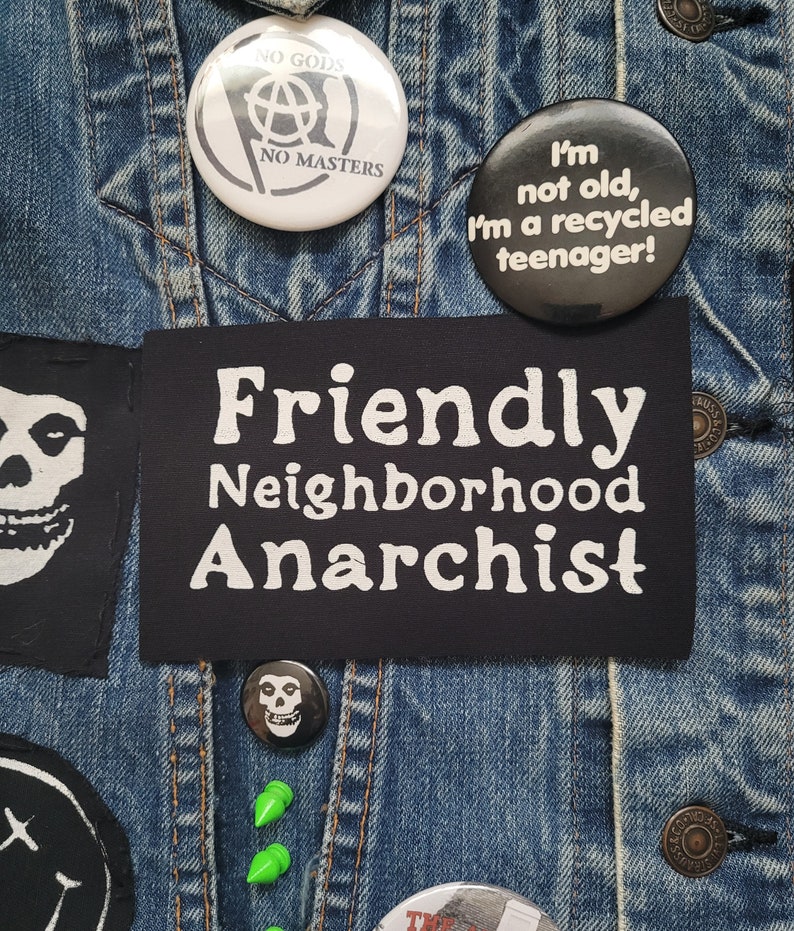 K&ouml;nnte beinhalten: Eine Jeansjacke mit verschiedenen Patches und Pins. Die Patches beinhalten einen schwarz-wei&szlig;en Totenkopf, einen schwarzen Patch mit wei&szlig;em Text, der "Friendly Neighborhood Anarchist" lautet, und einen schwarz-wei&szlig;en Pin mit einem Anarchiesymbol und dem Text "No Gods No Masters". Es gibt auch einen schwarzen Pin mit wei&szlig;em Text, der "Ich bin nicht alt, ich bin ein recycelter Teenager!" lautet.
