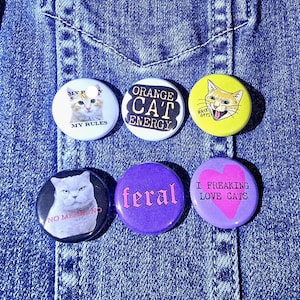 May include: Six round cat-themed buttons with various designs and text. The buttons feature images of cats, the words "Orange Cat Energy," "Feral," and "I Freaking Love Cats." The background is a blue denim fabric.