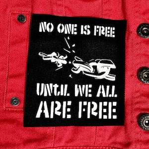 May include: A red denim jacket with a black patch. The patch features the text "NO ONE IS FREE UNTIL WE ALL ARE FREE" in white, along with a white graphic. The jacket has metal buttons.