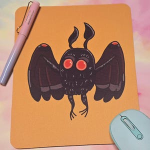 May include: A mustard-colored mousepad featuring a black moth-like creature with red eyes and wings. A light blue mouse and a pink and blue pen are also visible. The mousepad is rectangular and has rounded corners.