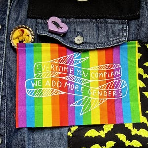 May include: A denim jacket with a rainbow patch that reads "Everytime You Complain We Add More Genders." The jacket also features a pin with a woman and a heart-shaped pin with a bat.