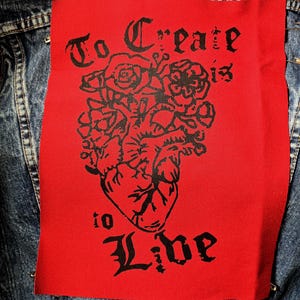 Könnte beinhalten: Ein rotes Stoffabzeichen mit den Worten "To Create is to Live" in schwarzer gotischer Schrift. Das Abzeichen zeigt eine schwarze Illustration eines Herzens, das mit Rosen und anderen floralen Elementen verschlungen ist. Das Abzeichen ist an einer Jeansjacke befestigt.