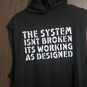 Puede incluir: Sudadera negra sin mangas con un estampado gráfico blanco que dice "El sistema no está roto, funciona como está diseñado."