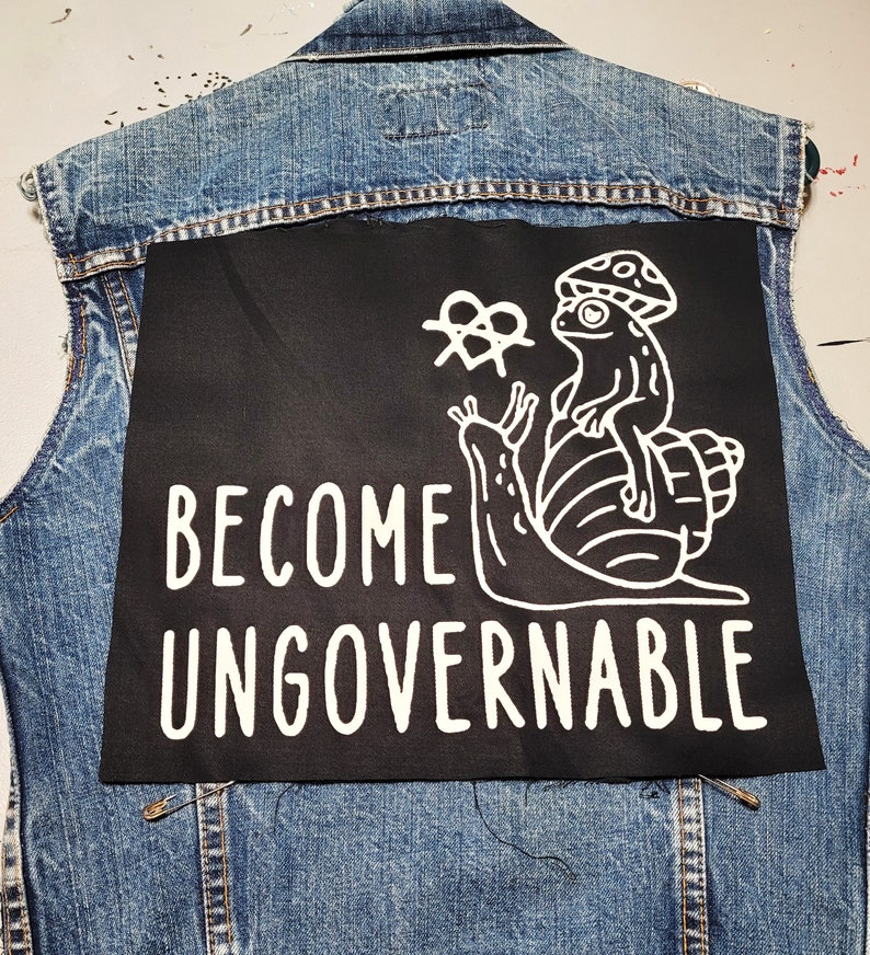 May include: A black denim vest with a black patch sewn on the back. The patch features a white illustration of a snail with a mushroom on its back and a frog sitting on top. The text "BECOME UNGOVERNABLE" is written in white below the illustration.