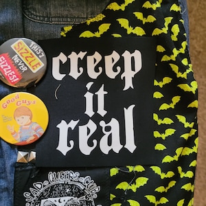 Op de afbeelding: Een zwarte patch met witte tekst die "creep it real" luidt, is genaaid op een spijkerjack. Het jack is versierd met andere patches, waaronder een gele patch met een cartoonfiguur en de tekst "Good Guys", een zwart-witte patch met een verdrietig gezicht en de tekst "Sad Queers Club", en een zwarte patch met een geel vleermuispatroon.