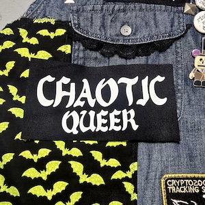 Peut inclure: Une veste en jean noire avec des patchs. Les patchs comprennent un patch noir avec du texte blanc qui dit "CHAOTIC QUEER", un patch jaune et noir avec des chauves-souris et un patch marron avec des bordures dorées qui dit "CRYPTOZOOL TRACKING SQUAD".
