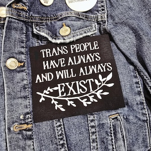 May include: Close-up of a denim jacket with gold buttons and a black patch. The patch reads "TRANS PEOPLE HAVE ALWAYS AND WILL ALWAYS EXIST" in white text. Two white buttons are also visible.
