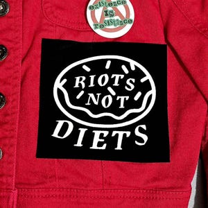 Pode incluir: Casaco de ganga vermelho com um patch preto com as palavras brancas "RIOTS NOT DIETS" num design em forma de donut. Acima do patch, um botão diz "existence IS resistance".