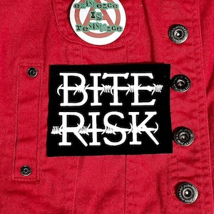 Op de afbeelding: Close-up van een rode spijkerjas met een zwart label met de witte tekst "BITE RISK" in een prikkeldraadlettertype. Boven het label staat een knoopspeld met de tekst "existence IS resistance". De jas heeft zilveren knopen.