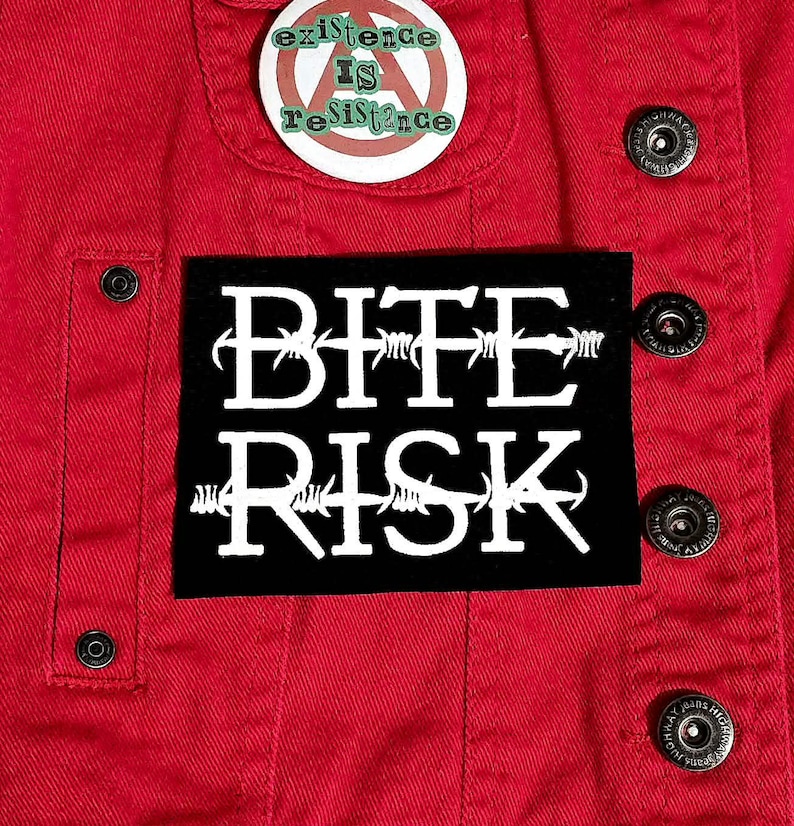 May include: A close-up of a red denim jacket featuring a black patch with the white text "BITE RISK" in a barbed wire font. Above the patch is a button pin that says "existence IS resistance". The jacket has silver buttons.