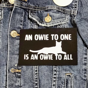 Może przedstawiać: Jeansowa kurtka z naszywkami i przypinkami. Czarna naszywka z napisem "AN OWIE TO ONE IS AN OWIE TO ALL" z białą sylwetką kota. Dodatkowe przypinki z napisami "NO WAR BUT CLASS WAR" i "WHEN INJUSTICE BECOMES LAW RESISTANCE BECOMES DUTY."