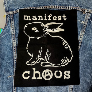 以下が含まれることがあります： 白いテキストと白いウサギのイラストが描かれた黒いパッチ。テキストは「manifest chaos」と書いてあり、「a」の代わりに無政府主義のシンボルが使われています。