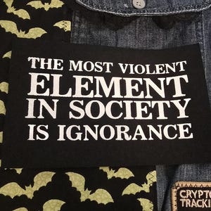 以下が含まれることがあります： 「THE MOST VIOLENT ELEMENT IN SOCIETY IS IGNORANCE」と書かれた白い文字の黒いパッチ。パッチは、黒いコウモリ柄の生地と他のパッチが付いたデニムジャケットにあります。