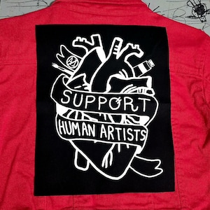 May include: Red denim jacket with a large black patch featuring a white anatomical heart design. The patch includes the text "SUPPORT HUMAN ARTISTS" in a banner, along with art supplies.