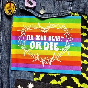 May include: A rainbow-striped patch with the text "FIX YOUR HEART OR DIE" in white, surrounded by a white barbed wire heart design. The patch is sewn onto a denim jacket, with other decorative pins and patches visible.